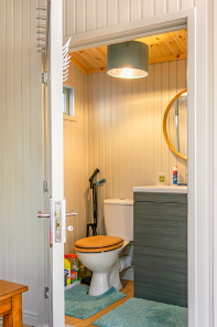 Open doorway leading into a compact bathroom with tongue-and-groove walls, toilet, vanity unit and wooden ceiling.